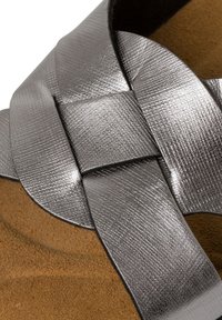 Cinturino della sandaletta in tono argento, testurizzato e con design intrecciato, abbinato a una morbida soletta in suede marrone per un contrasto elegante e comfort.