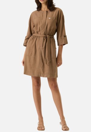 Femme portant une robe marron longueur genou avec manches trois-quarts, poches frontales et ceinture nouée, assortie à des sandales beiges à bouts ouverts.