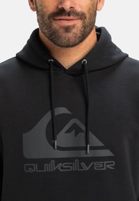 Fekete kapucnis pulóver zsinórokkal, elülső oldalon egy nagy, fényes szürke Quiksilver logóval és hegymintával. Puha, textúrált anyag.