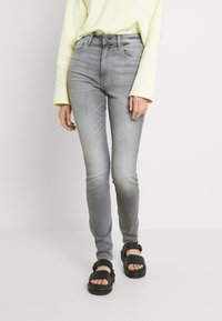Jeans gris de corte ajustado con una textura desgastada, una cintura de altura media y diseño de cinco bolsillos, combinados con sandalias negras. Top de manga larga de color amarillo brillante.