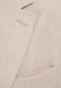 Beiger Fischgrät-Blazerstoff mit glatter Textur, ausgestattet mit einer einzelnen Tasche und dezenten Nähten am Revers.