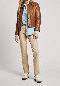 Giacca di pelle marrone con colletto rotondo, camicia azzurra chiara sotto, pantaloni beige e sneakers bianche con accenti marroni.