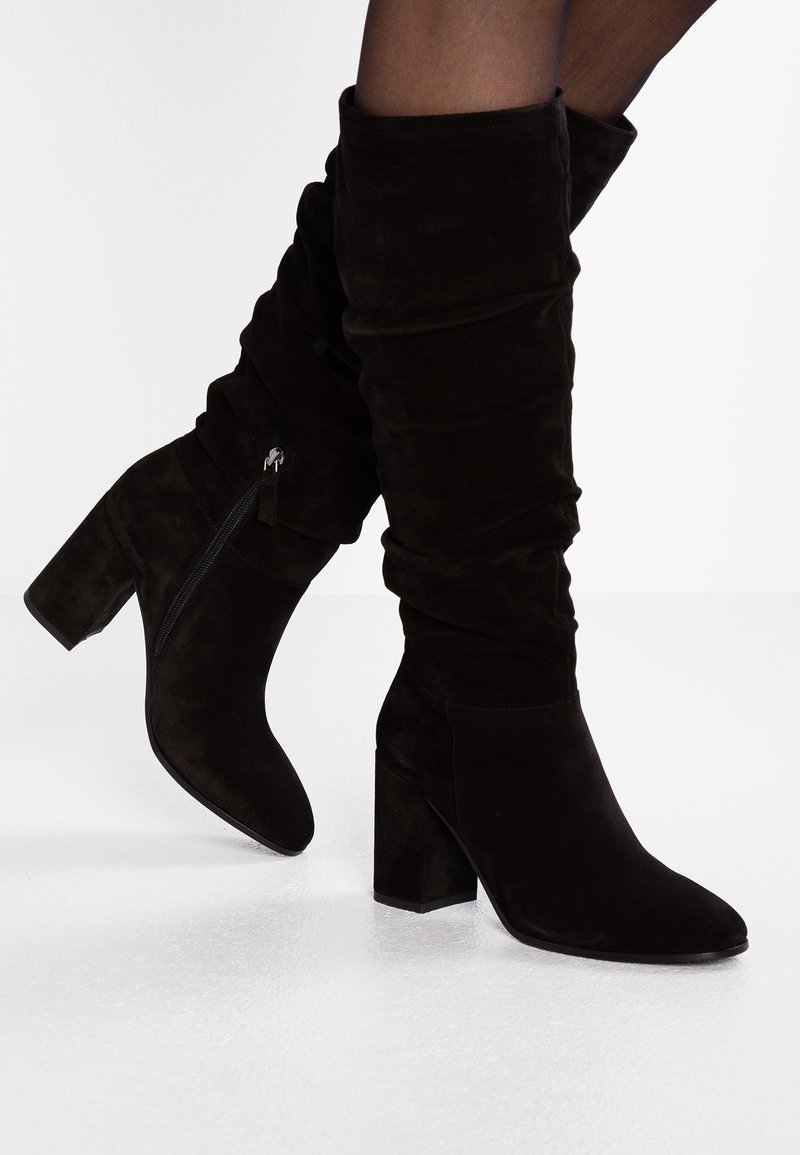 Svarta mocka knähöga stövlar med en staplad klack, som har en avslappnad design och en sidodragkedja för enkel påklädning. Slät textur och minimalistisk stil.