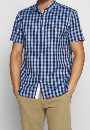 Mand iført en blå og sort ternet kortærmet skjorte med beige bukser, hænder i lommerne, foran en ensfarvet baggrund.