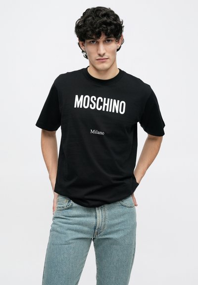 Juoda medvilninė marškinėliai su ryškiu baltu užrašu "MOSCHINO" ir žodžiu "Milano", trumpomis rankovėmis ir apvalia iškirpte. Dėvimi su šviesiai mėlynomis džinsais.