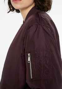 Burgunderfarbener Bomberjacke mit einer Reißverschlusstasche am linken Ärmel, geripptem Kragen und glatter, leicht glänzender Stoffbeschaffenheit.