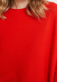 Czerwona sweter z dzianiny o teksturowanym wykończeniu, z okrągłym dekoltem i luźnym krojem. Materiał wydaje się miękki i przewiewny.