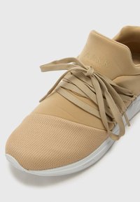 Pantofii sport bej prezintă un superior din plasă texturată, șireturi elastice și un design minimalist. Talpa este albă și are vârful rotunjit.