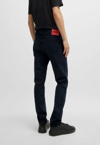 Jeans de corte ajustado en azul oscuro con una etiqueta de marca roja en la cintura. La tela tiene una textura suave. Se combinan con zapatillas negras.