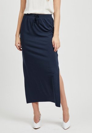 Woman wearing a navy blue ankle-length skirt with a side slit and elastic waistband, paired with white high-heeled shoes and a white top.