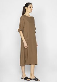 Brązowa, sięgająca do kolan sukienka z okrągłym dekoltem i krótkimi, falbaniastymi rękawami. Modelka nosi czarne sandały na płaskim obcasie. Gładka tekstura, swobodny styl.