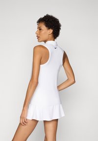 Kobieta z krótkimi kręconymi włosami, ubrana w białą tenisową sukienkę bez rękawów z małym czarnym logo z tyłu, stoi z jedną ręką na biodrze.