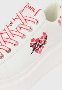Baskets blanches avec un motif floral rose et des lacets roses contrastants. Présente un logo noir brodé et des œillets argentés. Semelle épaisse blanche.