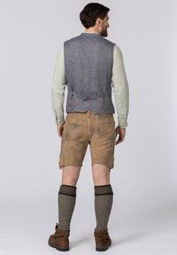 Graue Leinenweste über einem hellen langärmeligen Hemd, kombiniert mit braunen Lederhosen, knielangen grauen Kabelstricksocken und braunen Schuhen.
