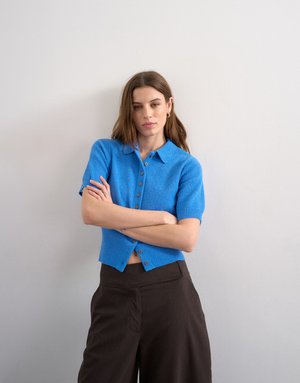 Barna hajú nő kék, rövid ujjú, gombolós kardigánban és sötétbarna nadrágban, keresztbe tett karokkal áll egy fehér fal előtt.