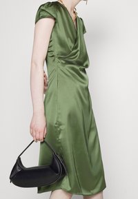 Robe en satin vert avec un décolleté drapé et de courtes manches en cap, accompagnée d'un sac à main noir aux formes incurvées et avec des surpiqûres contrastées.