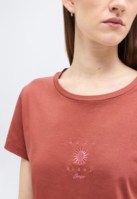 Γυναίκα που φοράει μπλουζάκι χρώματος σκουριάς με ροζ γραφικό ήλιου και κείμενο «AMAR EL SOL» κεντημένο στο στήθος.