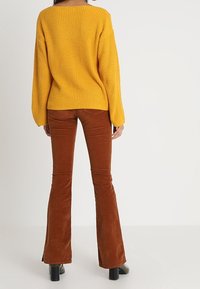 Żółty sweter z prążkowanego dzianiny zestawiony z brązowymi, rozszerzanymi spodniami z aksamitu. Sweter ma szeroki dekolt; spodnie mają gładką, lekko błyszczącą fakturę.