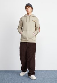 Felpa beige con cappuccio e logo NASA, tasca a marsupio e dettagli rosa. Abbinata a pantaloni larghi in velluto a coste marroni e sneakers bianche.