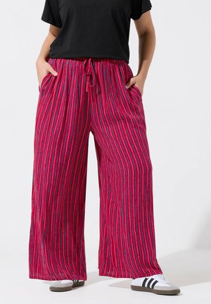 Personne portant un pantalon rouge à jambes larges avec des rayures verticales bleues et blanches, associé à des baskets blanches et un haut noir à manches courtes.