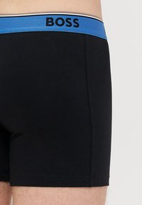 Μαύρα μπόξερ από μαλακό βαμβάκι, με μπλε ελαστική μέση που φέρει το όνομα της μάρκας "BOSS" σε μαύρα γράμματα.