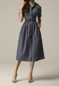 Kvinna som bär en knälång, kortärmad, knäppt grå klänning med ett svart bälte och nakna högklackade sandaler mot en enkel bakgrund.