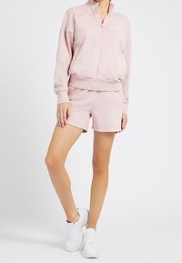 Rosa Zip-Up Sweatshirt mit hohem Kragen, kombiniert mit passender Shorts. Beide bestehen aus weichem Stoff, verfügen über ein Logo-Detail und haben eine lässige Passform.