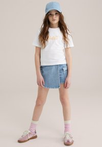 Camiseta blanca con "Barbie" bordado en naranja, acompañada de una falda envolvente de mezclilla, gorra azul claro, zapatos rosas y calcetines a rayas.
