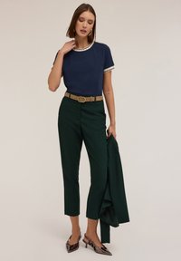 Tricou bleumarin cu margini albe, pantaloni verzi cu talie înaltă, curea bej și pantofi negri lustruiți cu toc. Aspect croitorit, ajustat.
