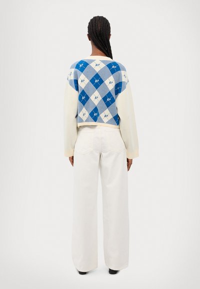 Pull en crochet bleu et blanc à motif losanges, avec des manches longues beiges, associé à un pantalon large blanc et des chaussures noires.