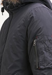 Musta vuorattu takki, jossa on karvavuorattu huppu, vasemmassa hihassa vetoketjullinen tasku ja ommeltuja yksityiskohtia kankaassa.