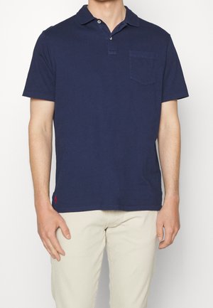 Mężczyzna w granatowej koszulce polo z krótkim rękawem i kieszenią z przodu oraz beżowych spodniach, stojący na jednolitym białym tle.