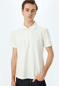 Krämfärgad kortärmad polo med dragkedja i kragen, tillverkad av ett slätt tyg, med en avslappnad passform och diskreta sömdetaljer.