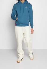 Persona che indossa una felpa con cappuccio blu Alpha Industries, pantaloni texturizzati color crema e sneaker beige, in piedi su uno sfondo chiaro e semplice.