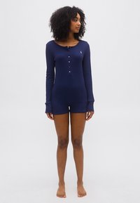 Γυναίκα με σγουρά μαλλιά, ντυμένη με μακρυμάνικο ρούχο τύπου ρομπέρ σε ναυτικό μπλε, όρθια ξυπόλυτη σε απλό φωτεινό φόντο.