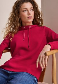 Roter Kapuzenpullover mit Kordelzug und langen Ärmeln. Weicher Stoff, lockere Passform, getragen mit blauen Jeans, sitzend auf einem Holzstuhl.