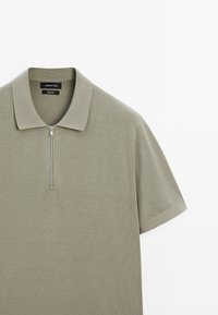 Polo à manches courtes vert olive clair avec un col et une fermeture éclair argentée à l'avant, en tissu tricoté 100 % coton.