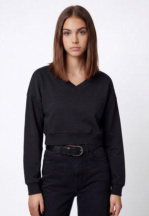 Ung kvinde med lige brunt hår iført en sort V-hals cropped sweatshirt og højtaljede sorte jeans med sort bælte.
