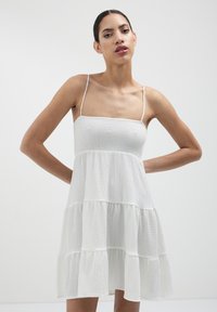 Abito estivo bianco a strati con spalline sottili, tessuto strutturato e busto aderente. Presenta una vestibilità comoda con strati arricciati all'orlo.