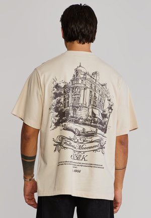 Uomo che indossa una maglietta beige con un disegno dettagliato nero di un'auto d'epoca e un edificio storico accompagnati da testo decorativo sul retro.