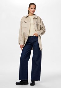Beige oversized skjortjacka med två bröstfickor, mörkblå vida jeans och svarta klumpiga stövlar. Slät textur och neutrala färger.