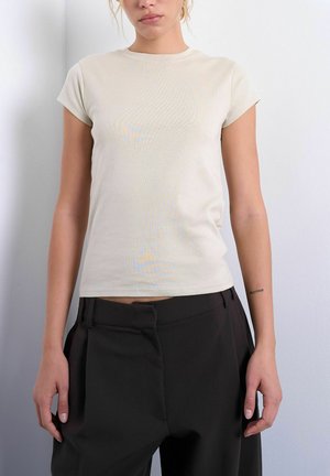 Woman wearing a fitted beige short-sleeve top and loose black high-waist pants standing against a light wall.