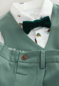 Gilet vert avec une chemise blanche ornée d'un motif animalier coloré et un nœud papillon en velours vert foncé. Comprend un détail de bouton marron.