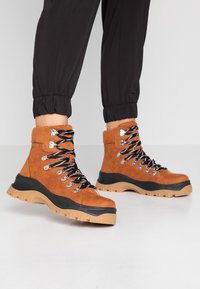 Bottes de randonnée en daim marron avec des lacets noirs, des œillets métalliques et une semelle épaisse beige. Le design comprend une cheville rembourrée pour un meilleur maintien.