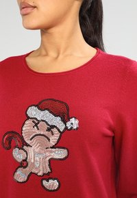 Femme portant un pull rouge avec un bonhomme en pain d'épice triste à paillettes, coiffé d'un chapeau de Père Noël et tenant une canne en sucre d'orge.
