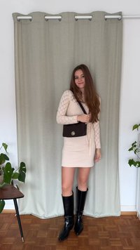 Crèmewitte, geribbelde gebreide jurk met lange mouwen, gecombineerd met kokerhoge zwarte leren laarzen en een kleine zwarte crossbodytas. Zachte achtergrond en planten.