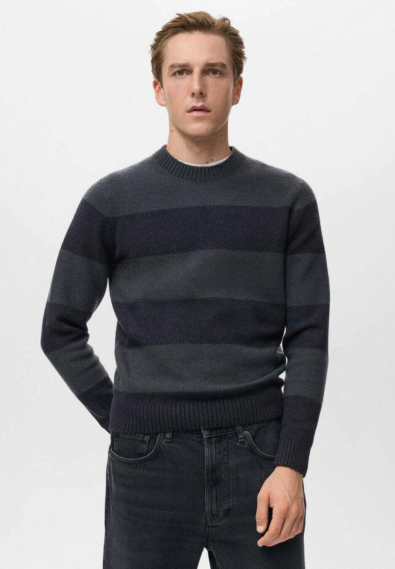 Giovane in piedi, che indossa un maglione a righe orizzontali grigio scuro e nero e jeans neri, su uno sfondo chiaro e uniforme.