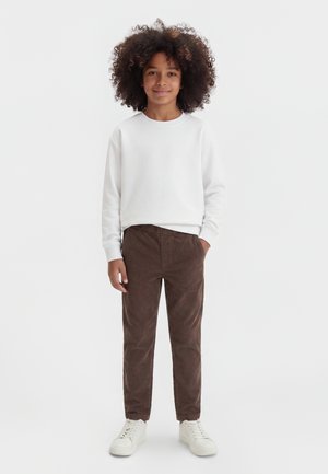 Enfant aux cheveux bouclés portant une chemise blanche à manches longues, un pantalon marron et des baskets blanches, debout avec une main dans la poche.