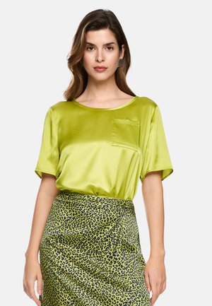 Woman wearing a shiny lime green short-sleeve blouse with chest pocket and high-waisted green and black leopard print skirt.