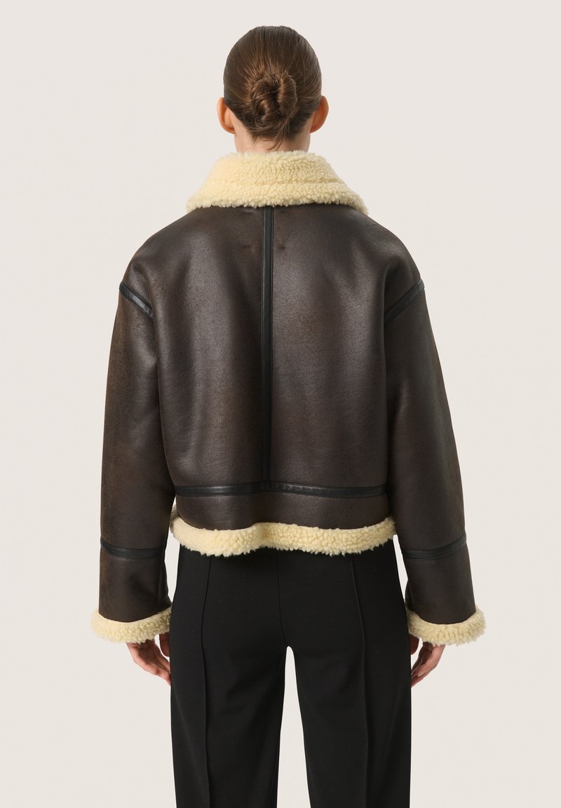 Giacca in shearling marrone con collare e polsini in shearling beige. Presenta un design cropped e cuciture verticali sulla schiena. Texture liscia.